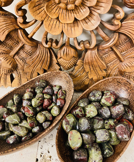 Two bowls containing tumbled Dragons Blood Jasper stones, with varying colors and patterns, displayed in front of a wooden decorative panel.