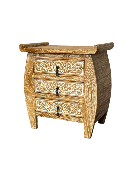 A white washed teak wood nightstand with three drawers, featuring intricate carvings on the front and a curved design at the top.